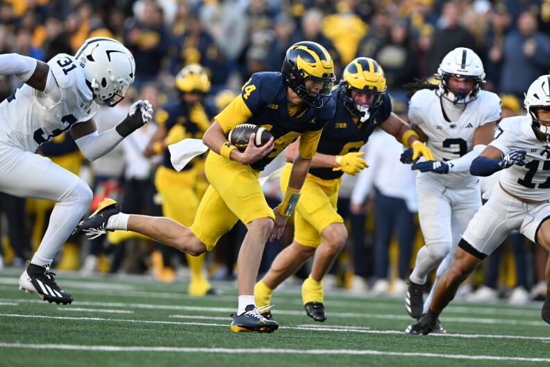 Michigan quarterback Tommy Carr (14) in a maize-and-blue uniform runs with the football as white-clad defenders close in during an intense college football game.