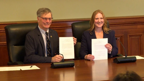 A grey haired man with glasses and a suit holds up a piece of paper. Next to him a blond haired woman holds up a piece of paper. 
