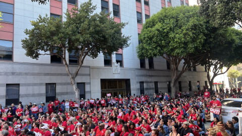 A crowd of more than a thousand nurses wearing red rally in front of a large building with grey cinderblocks and verticle windows, blocking the entire street and sidewalk. 