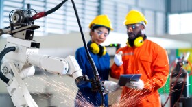 Two people in bright-colored suits watch a robot arm as it welds.