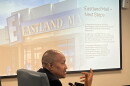 In the foreground, a man addresses someone offscreen. A slideshow is displayed behind him 