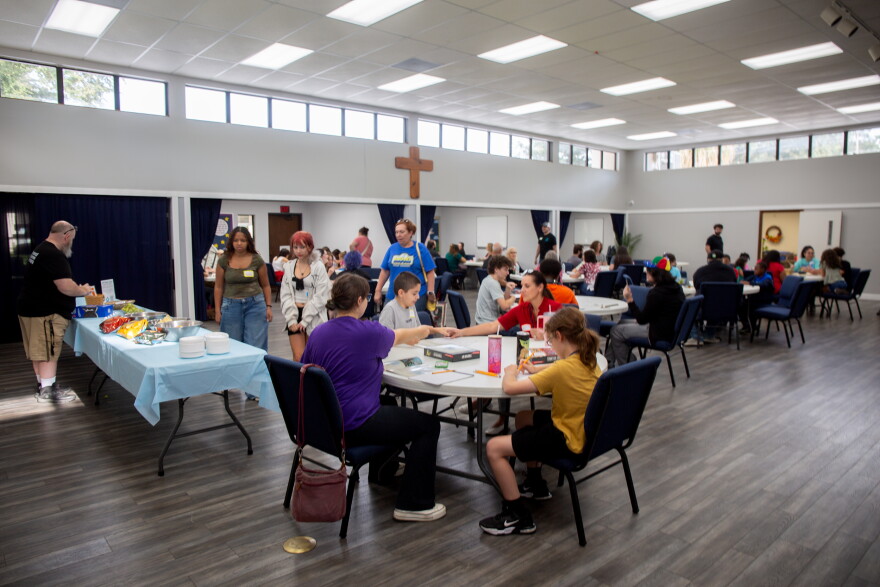 A large event space inside a church with wood laminate flooring and white walls. The room is filled with folding circular tables filled with people playing board games. 
