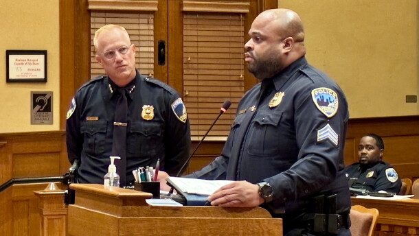 A police officer stands at a podium speaking, while another officer stands behind him and a third officer sits at a desk in the background.