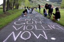 High school students write chalk messages on a sidewalk in a grassy area. The most prominent one reads "END GUN VIOLENCE NOW."