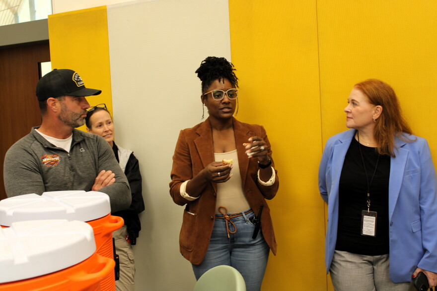 Three people stand in a room with yellow wall panels while a woman in a brown blazer speaks and holds a small item.