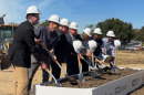 Seven people wearing white construction hats and holding shovels stand in front of a raised box of soil