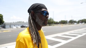  A Black man with long hair and a beard stands in a yellow shirt in front of a crosswalk.