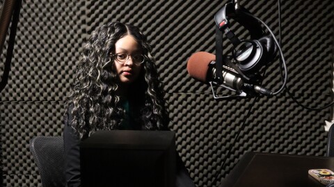 A woman with long curly hair and glasses sits in a soundproof radio studio, facing a microphone.