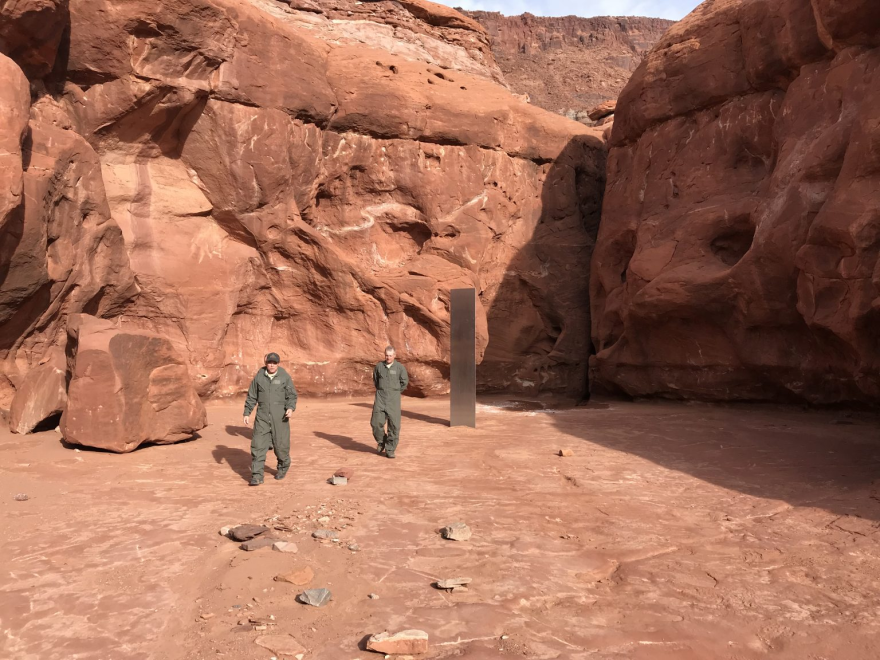 A photo of two men in green suits next to the silver-colored monolith. 