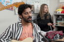 Close-up of Rafay Rashid playing guitar and singing. He's wearing a black and white striped shirt over a peach t-shirt. Talia Temporal plays a drums in the background.