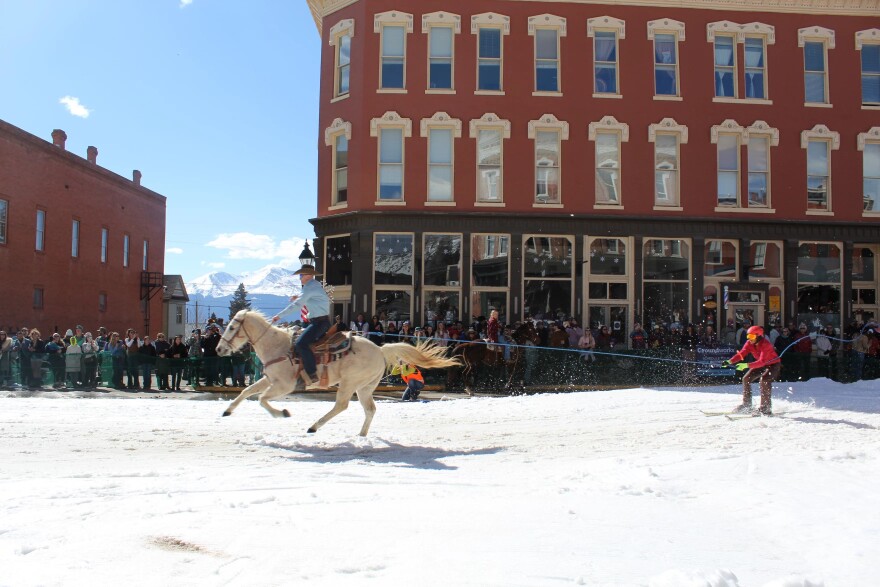 The Sawatch Range, including fourteener Mt. Massive, looms in the background as a cowboy tows a skier down Harrison Avenue during Leadville Skijoring in March, 2025.