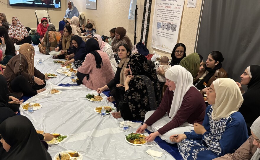 A group of women sit on blue and white tarpulin sheets with plates of food and soda. They are all wearing headscarves.