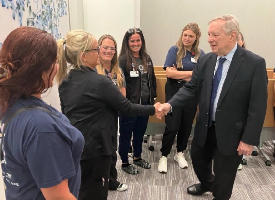 A man in a suit shakes hands with a woman among a group of smiling people, some wearing medical scrubs, standing indoors in a well-lit room with white walls and a carpeted floor.