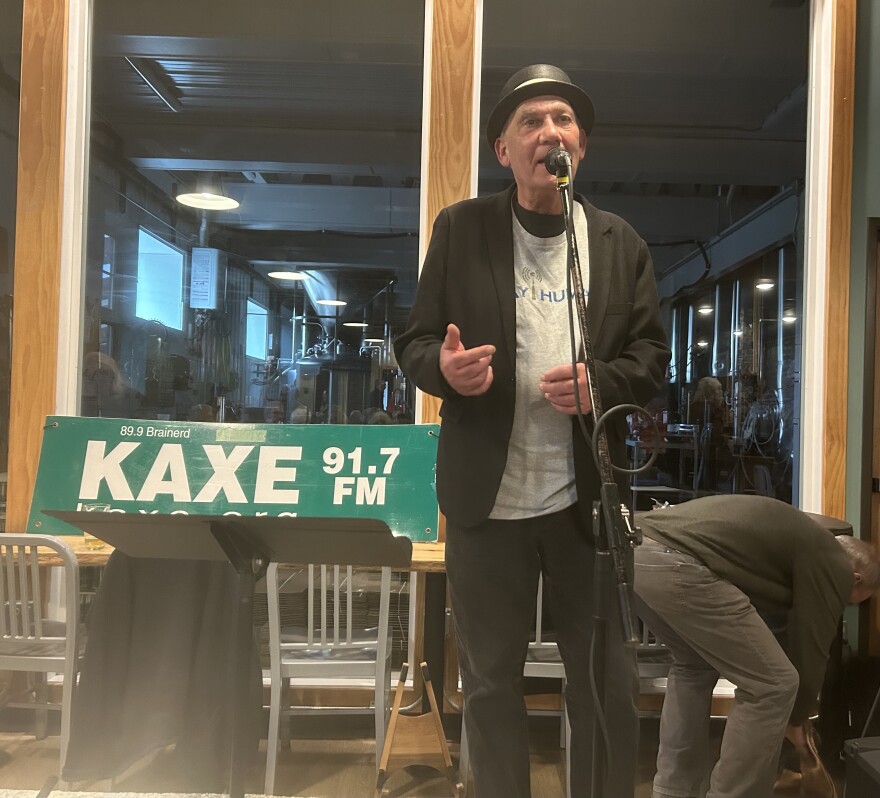 A man standing on stage wearing a hat with a green sign behind him that says KAXE 91.7 FM