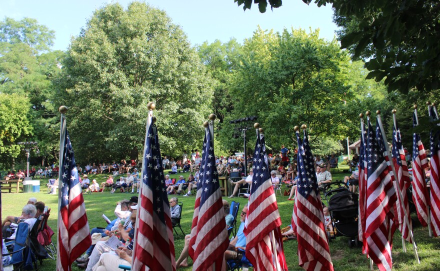 American flags stand in the foreground as a crowd sits on the lawn watching the program near the amphitheater.