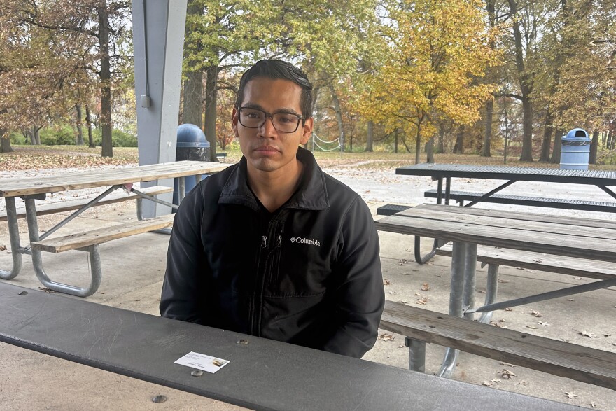 Juan Carlos Valladares-Hernandez sits at a picnic table under a shelter in a park. He's wearing a black jacket and his shoulders are slumped. Behind him are trees with leaves turning yellow.