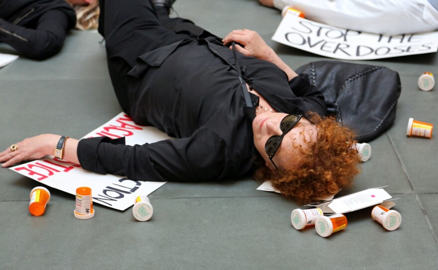 A protestor in sunglasses and black clothing lies on the ground next to empty pill bottles and a sign that reads "STOP THE OVERDOSES."