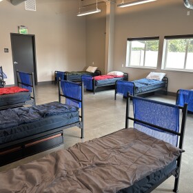 Eight single beds are lined up in a room with a concrete floor and windows. 
