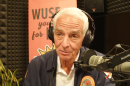 man with white hair is wearing headphones in a radio studio talking into a microphone. he is wearing black jacket over a white button shirt. the jacket has a round logo on it 