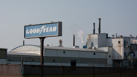 The Goodyear chemical plant is pictured. In front of it is a Goodyear logo sign, which is blue with white text that reads "Good," then an image of a stepping shoe with wings, and then the word "year." The factory behind it has a dark wire fence surrounding it, and is white and grey. The factory has a white puff of smoke coming out of the top of it. Rust runs down the side of some of the outer walls.