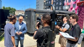 A three-person television crew films a host in sunglasses and a blue blazer interviewing a man with long hair and a beard in a food-truck park.