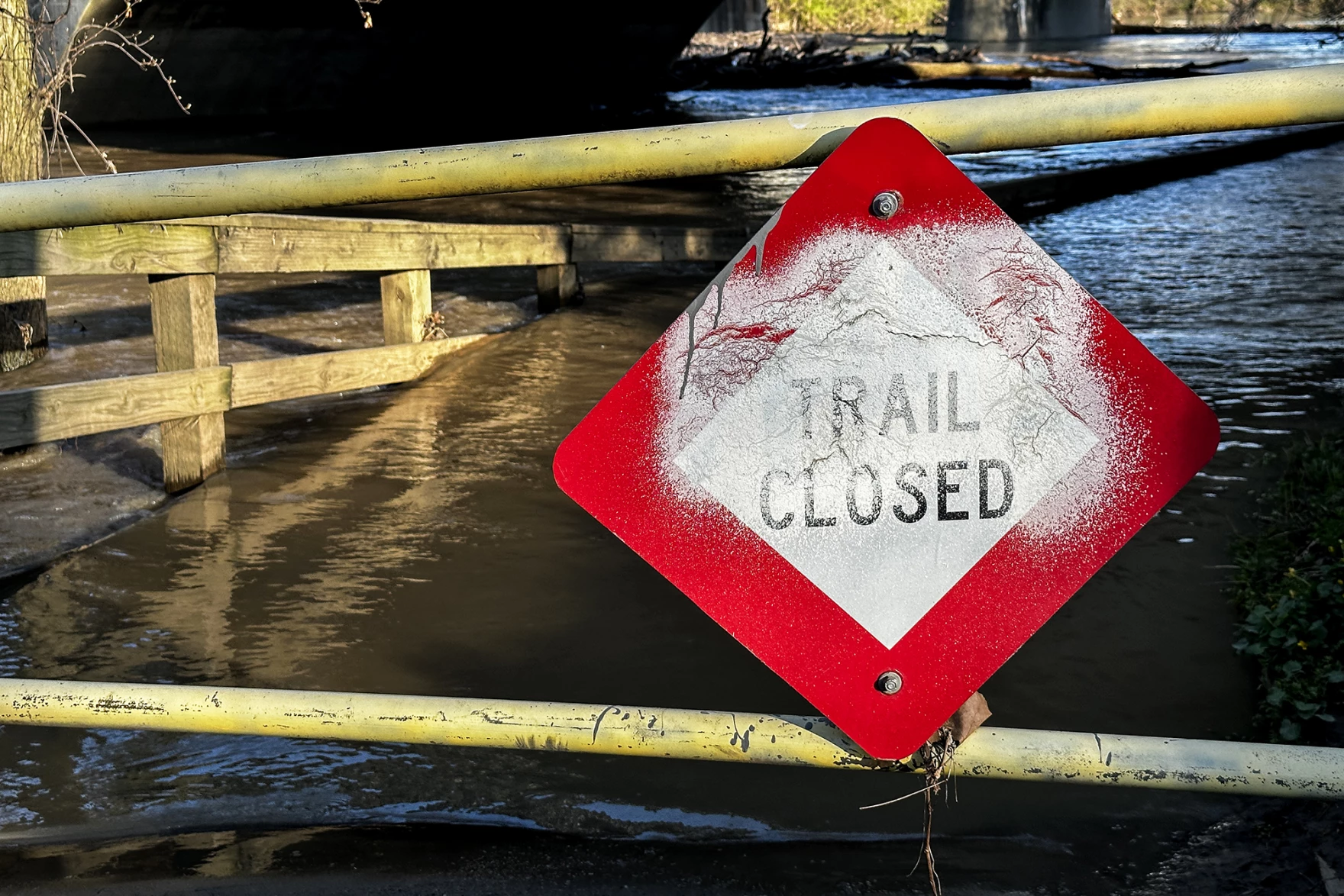 A sign on a trail that reads 'Trail Closed.' Behind the sign flood waters creep up the path.