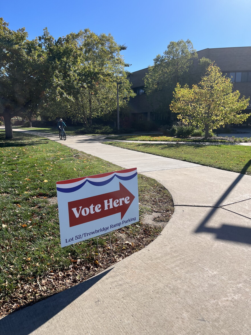 A red, white and blue Vote Here sign beside a campus sidewalk, with trees and a cyclist in the background.