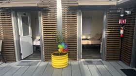 Two tiny home units with brown paneled exteriors have their doors open, showing beds inside. A yellow plant pot is outside between the two units.