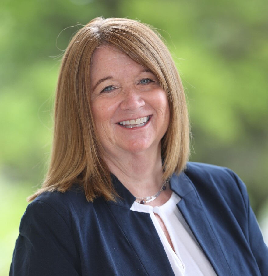 A woman with shoulder-length strawberry blonde hair and blue eyes is smiling. She is wearing a blue blazer with a white shirt underneath, in addition to a silver necklace with a few black beads.