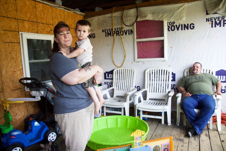 A woman holds a young boy on an unfinished porch while a man looks on.