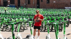 A man in a red shirt and black shorts stands amidst hundreds of lime-green bikes and scooters, looking at his phone and typing.