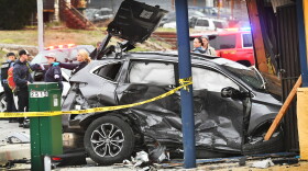 A crumpled, gray SUV sits smashed into the side of a building. First responders can be seen in the background working and emergency vehicles are in the background with lights flashing.