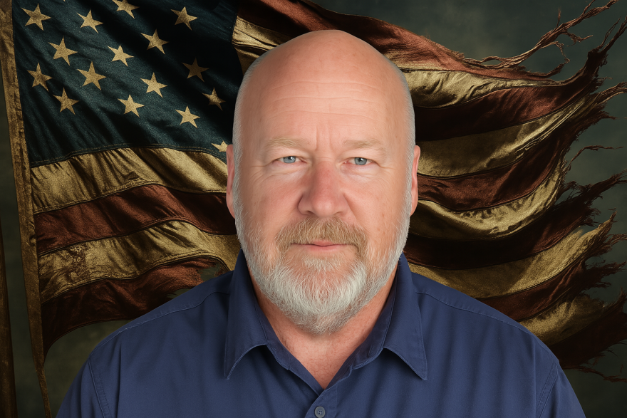 A man wearing a dark blue collared button-up shirt looks into the camera. A tattered American flag waves in the background.