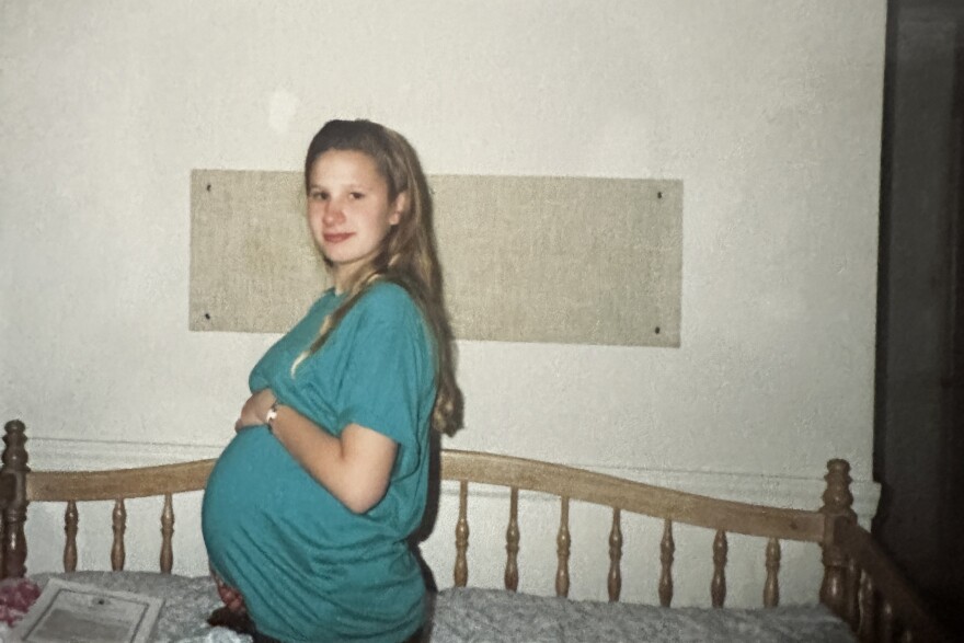 A 1991 photo shows St. Louis native Toni Popham standing by her bed in the Godparent Home in 1991. Pregnant and 13, she joined other girls living in the maternity home for months before delivery. She is wearing a green or teal short sleeved tshirt, and cradles her belly with both hands. In a new podcast, Popham and others are speaking out about the pressure they faced to give their babies up for adoption.
