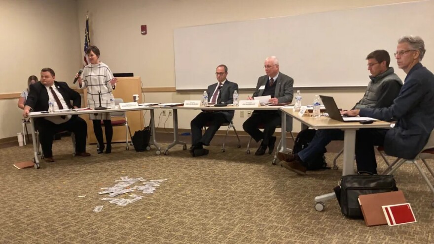 Ryan Richardson stands holding a microphone behind a row of tables. Five men are seated at the tables. A pile of play money is on the floor in front of them.