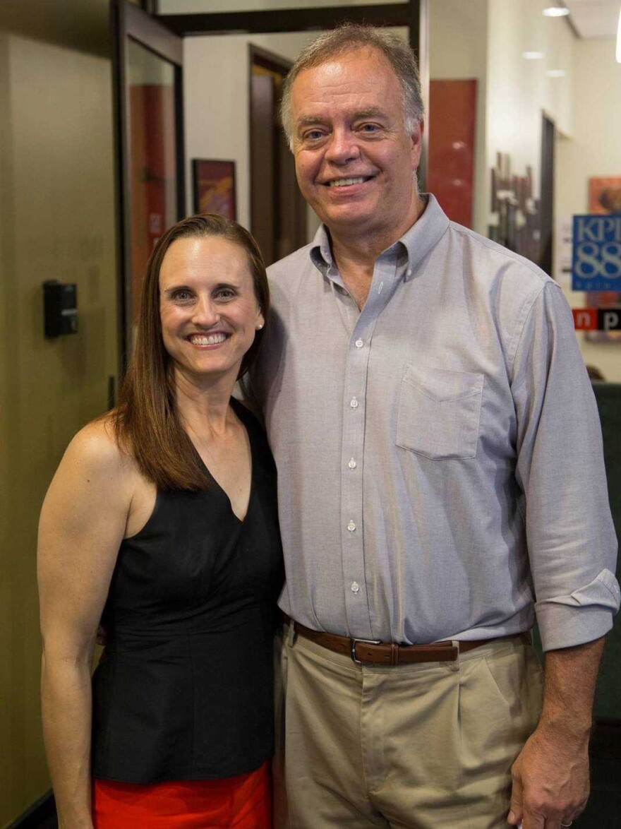 A woman in a black top and a man in a light blue button-down shirt pose in an office space.