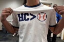 A man grabs the sides of his white shirt to showcase the text that reads "HC > Baseball," but the baseball is an icon, and not words.