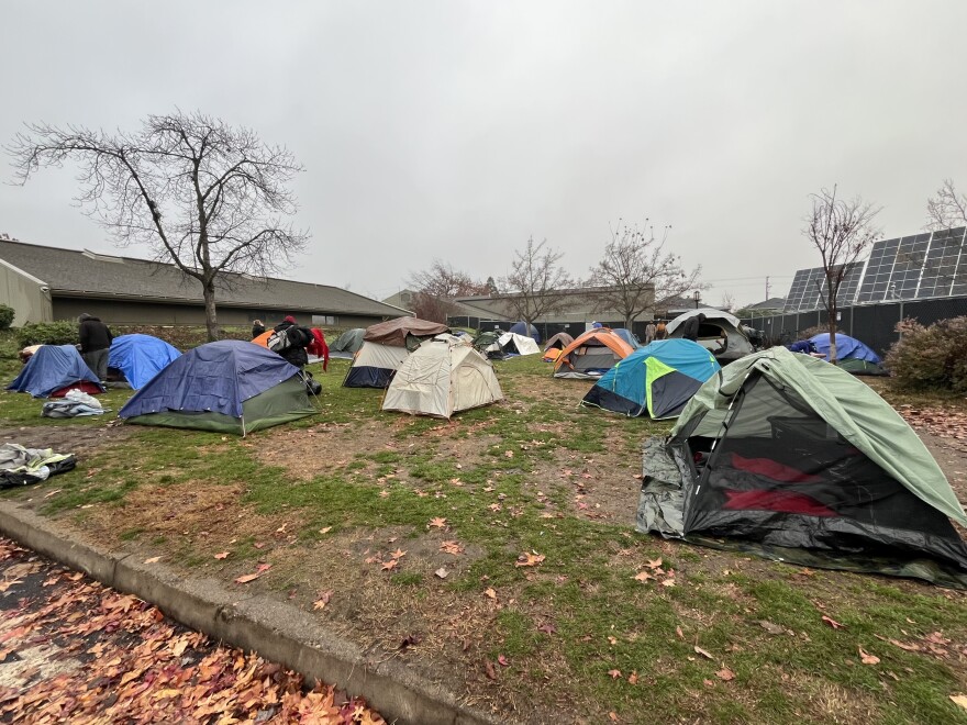 Colorful tents and tarps are set up on a grassy lot. The area is surrounded by buildings and solar panels. 