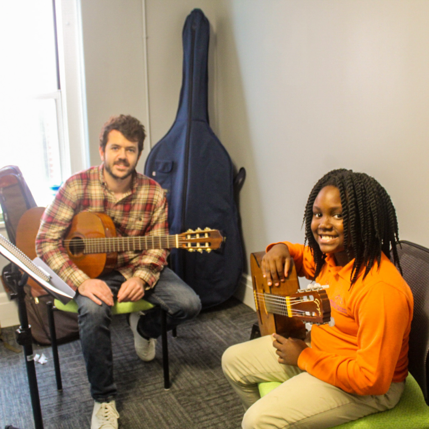 A picture of a young person in long black dreads and orange shirt holding a guitar and smiling. In the background is another person in a plaid shirt, they are holding a guitar and smiling. 