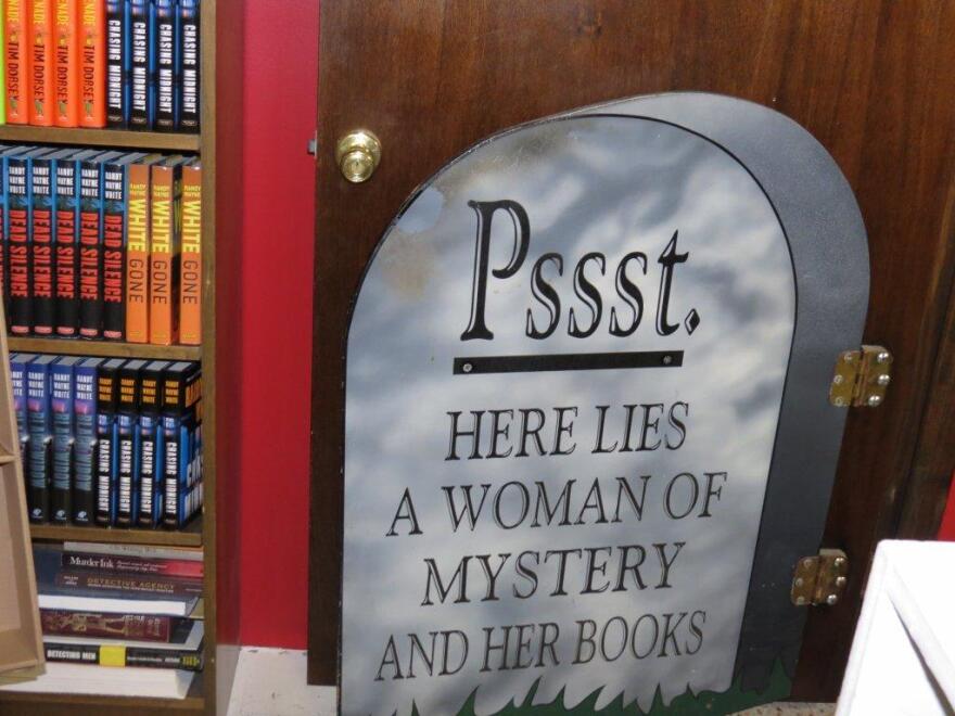 A view of the stacks in the Murder On the Beach Mystery Bookstore. A sign depicting a tombstone reads 'Psst. Here Lies A Woman Of Mystery And Her Books'