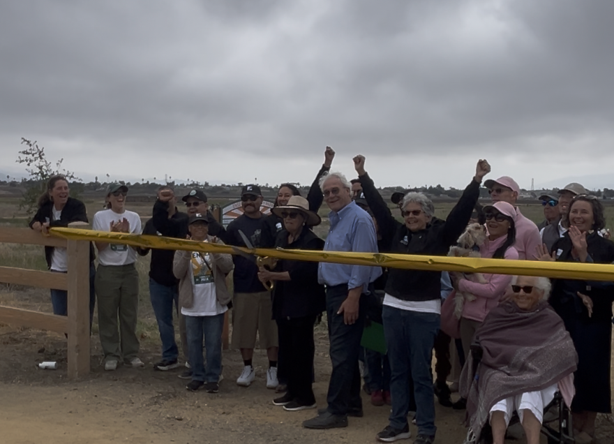 A woman with large scissors poised to cut a golden ribbon extended in front of a crowd of people. Two people have their hands raised. Others are clapping and waving.