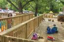 A large wooden deck is pictured at the back of tiger tots. The deck is attached to two of the buildings and overlooks the parking lot and a few trees. The deck is filled with toys of all colors and chairs for teachers and supervisors.