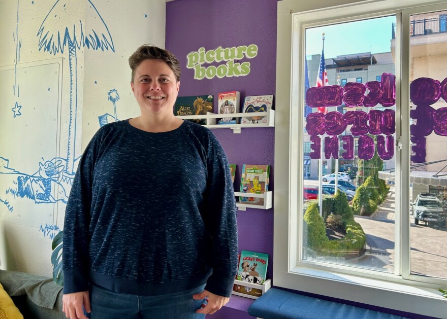 A woman stands next to a window, in front of a wall decorated with a comic illustration and some picture books on display.