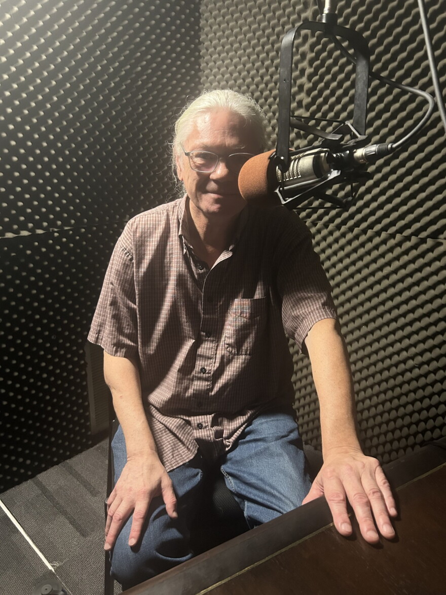 A man with long grey hair tied into a ponytail  and glasses sits behind the microphone in a studio with padded walls.
