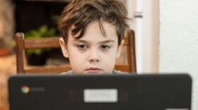 A child sits behind a laptop, looking at the screen. 