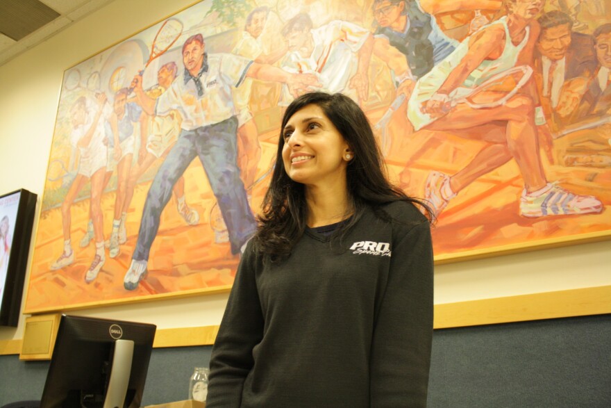 Shabana Khan stands in front of a painting at Pro Sports, Bellevue, that depicts members of her family playing squash. (Shabana is the one in blue, while her brother Azam is in the center of the painting at her left).