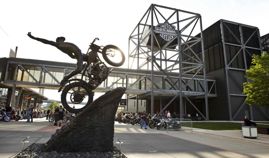A sculpture of a person riding a motorcycle up a steep hill with several rows of motorcycles in the background.