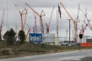 Large, metal cranes rise above an entrance to a construction site. A sign, partially obscured by a chain-link fence, reads, "Intel." 