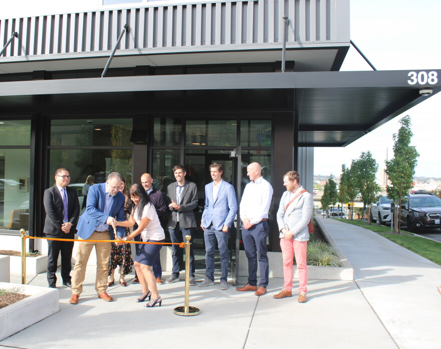Project leaders stand in front of a new gray and white apartment complex, cutting a ribbon in front of an open entrance. 