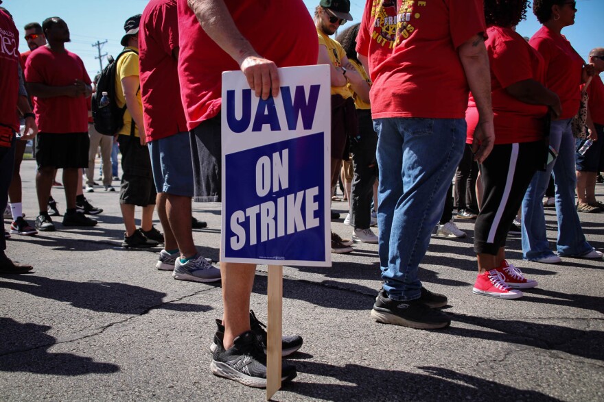 A man holds a sign by his leg that reads "UAW on strike" many stand around him